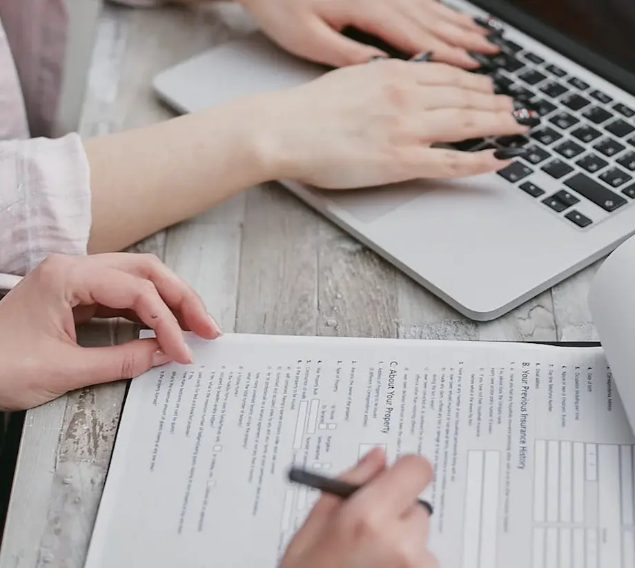 Person filling out a form on paper while another types on a laptop keyboard.