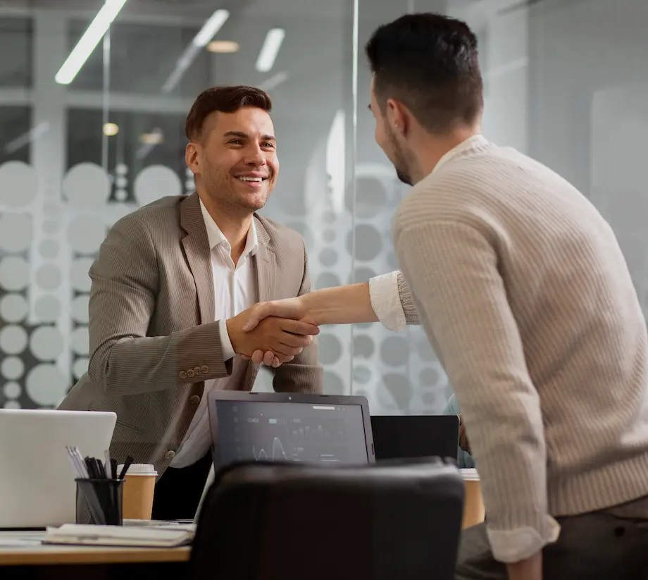 Dwóch mężczyzn podających sobie ręce przy biurku w nowoczesnym biurze, co sugeruje udane spotkanie lub zawarcie umowy.